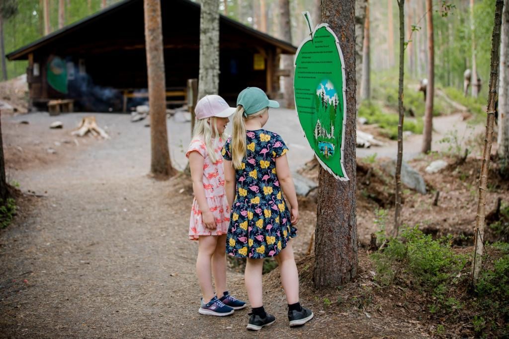 Lapset lukevat lehdenmuotoista kylttiä Laavun edustalla.