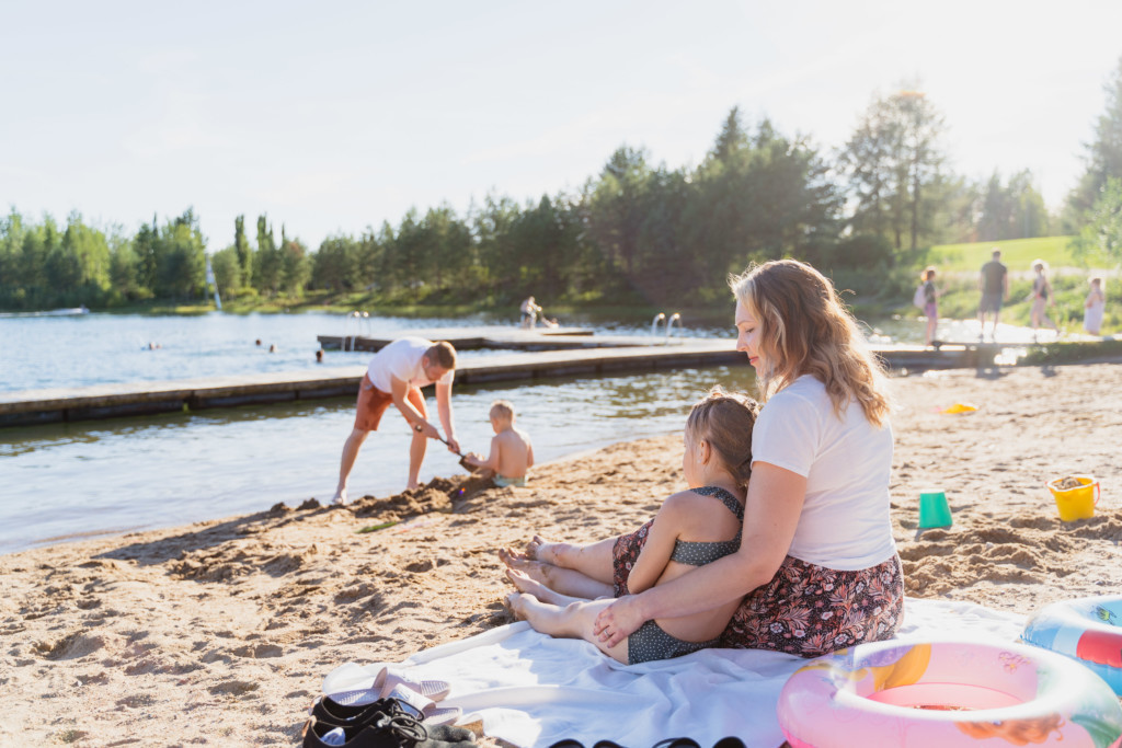 Perhe uimarannalla, nainen pitelee tytärtä kainalossa, isä ja poika leikkivät kauempana hiekalla